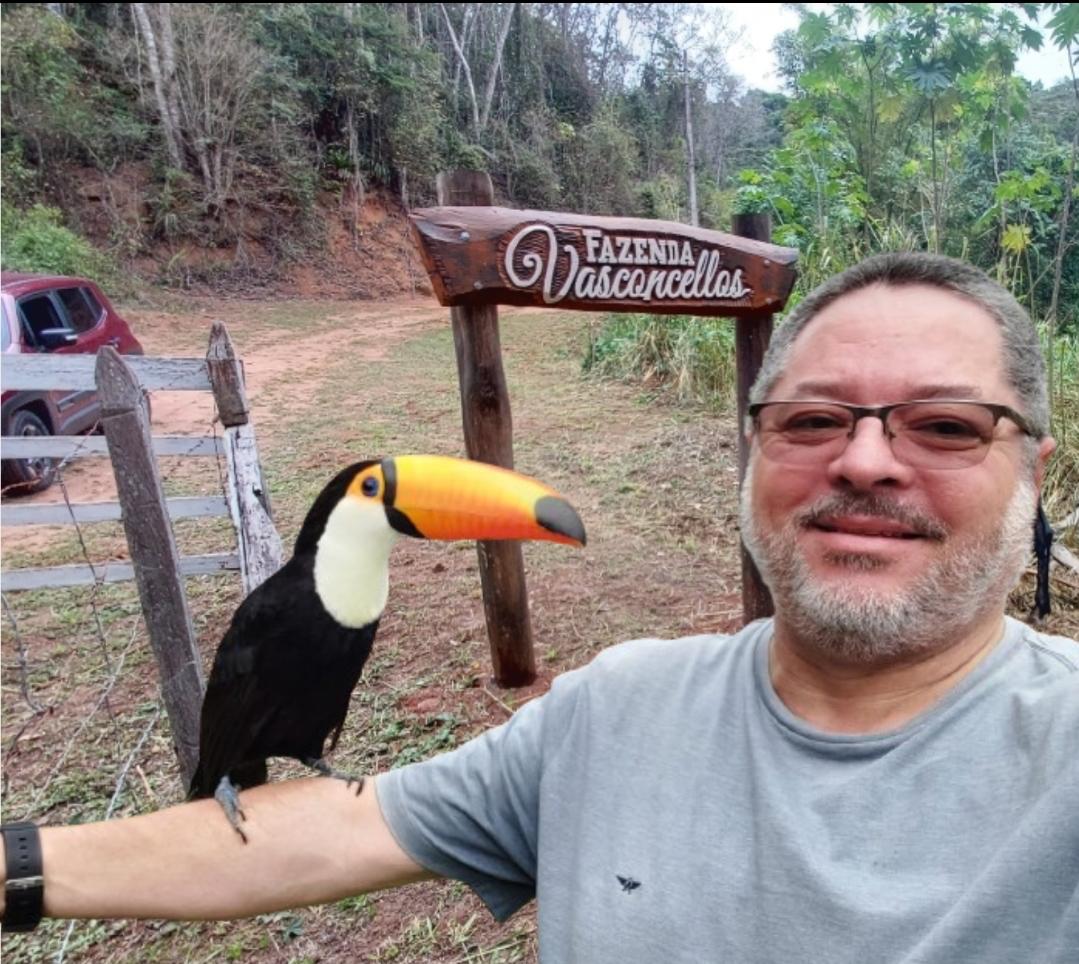 Homem sorrindo com um tucano pousado no braço junto ao portal da fazenda